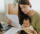 Mother sitting with baby in her lap, using laptop to check finances at home.