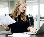 Businesswoman with long blond hair holding document and using laptop in modern office