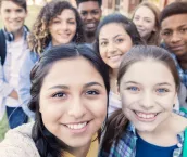 Diverse group of eight high school students are smiling and looking at the camera. Teenagers are students at public high school, and are wearing backpacks or holding school books.