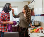 Mature woman is cooking a curry in the kitchen of her home. She is feeding her son a piece of sliced pepper as he does the dishes.