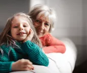 A girl and her grandma sitting on a sofa and smiling at the camera