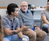 Mid adult male army veteran has an intense conversation with an unrecognizable person during a support group meeting. He gestures while speaking.