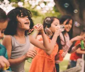 Long haired boy laughing and having fun with his friends standing on a wooden fence in a summer park blowing bubbles, with a vintage develop