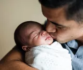 A father gently kisses his newborn daughter on the cheek. He is holding her cradled in his arms as she is swaddled up comfortably for her nap.