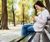Young woman sitting on the bench in public park smiling and checking social media or messages on smartphone. Young woman sitting on the bench in public park smiling and checking social media or messages on smartphone.