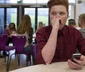 Stressed schoolboy sitting away from people at school. He has a smartphone in his hand and is looking away with his hand to his face.