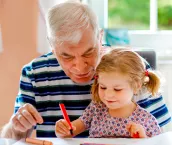 A senior man and his granddaughter are sitting on at a table colouring on a piece of paper. A senior man and his granddaughter are sitting on at a table colouring on a piece of paper.