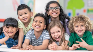 Portrait of a happy multi-ethnic group of kindergarten age students. The cute children are laying in a pile on the ground in a modern classroom. The kids are laughing and smiling.