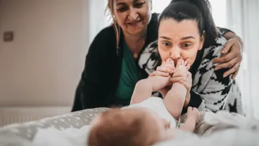 A young mother lovingly kisses the feet of her infant baby as her mother hugs her and watches from behind.