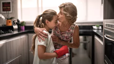An older woman in the kitchen leans down to kiss a young girl on the forehead. An older woman in the kitchen leans down to kiss a young girl on the forehead.