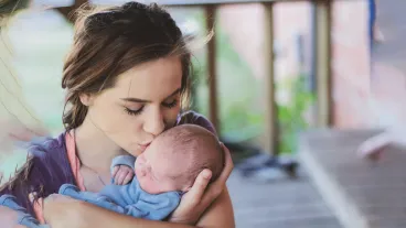 Young teenage woman is mother to newborn baby boy. She kisses him on the cheek as she holds him lovingly in her arms.