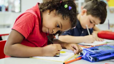 Close-up of young Hispanic schoolgirl sitting at classroom table and concentrating on drawing with colored pencils.