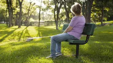 Rear view of one mature woman sitting on a park bench in the summer season. Bright sunny day with lush green grass and trees. The woman gazes off into the distance as she relaxes on a beautiful day. Solitude, lonliness, contemplation. She has short blond hair and wears a purple shirt and jeans. Copyspace to right in this tranquil nature scene. Rear view of one mature woman sitting on a park bench in the summer season. Bright sunny day with lush green grass and trees. The woman gazes off into the distance as she relaxes on a beautiful day. Solitude, lonliness, contemplation. She has short blond hair and wears a purple shirt and jeans. Copyspace to right in this tranquil nature scene.