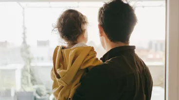 Rear view of dad holding his baby boy looking out of the window at home.