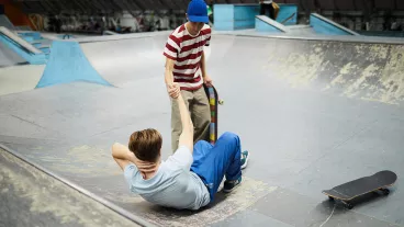 Two teenage boys are in a skate park. One boy is laying on the ground on his back, he has fallen. The other has extended his arm to help him back on his feet. 
