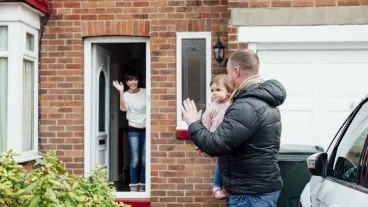 Father is carrying his baby daughter to the car and is turning round to wave at the mother who is stood at the front door of the house.