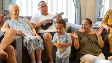 Pacific Islander family sitting together at home. The toddler is playing a ukulele and they are all clapping.