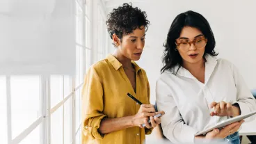 Two business women having a discussion, they're standing in an office and using a tablet