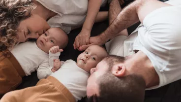 Happy young family with newborn twin babies lying on the bed together, parents holding hands and kissing their babies