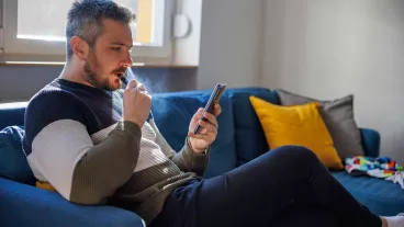 Man sitting on the sofa at home and using smart phone for home finances, smoking electronic cigarette and looking serious