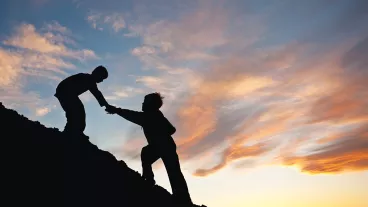 Silhouette image of two boys climbing a hill with one reaching out to help the other. 