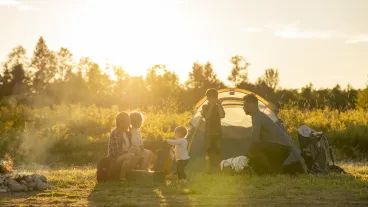 A small family settles into the campsite they have just finished setting up as they prepare to enjoy a vacation together. They are each dressed comfortably and some are finishing placing items in the tent while other enjoy sitting by the fire and relaxing