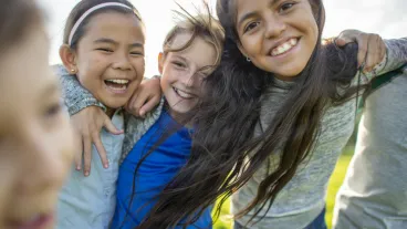 A small group of Elementary children huddle in closely to one another as they pose for a portrait. They are each dressed casually and are smiling as they spend the time together.