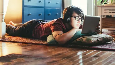 Teenager attending online classes at home during lockdown caused by COVID-19 world pandemic. He is looking at digital tablet with headset on his head while on the floor of his living room.