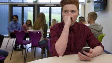 Stressed schoolboy sitting away from people at school. He has a smartphone in his hand and is looking away with his hand to his face.