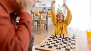 A delighted granddaughter raises her arms in celebration after winning a game of chess at home. Her face lights up with joy and pride as she revels in her success, while the peaceful home setting adds to the warmth of this special moment of achievement.