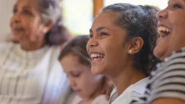 Multi generational women on the sofa. The young girls are sisters. One is a teenager on is younger. There is also a mother and grandmother. They are all happy and smiling. They are all Australian Aboriginals.