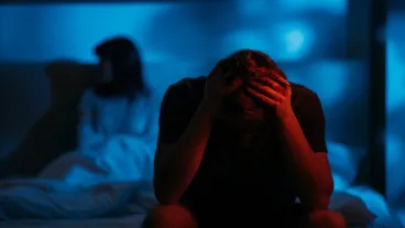 Depressed devastated man clutching head sitting on bed on distance from offended wife in dark red blue neon light on defocused background.
