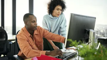 Waist-up view of casually dressed Black and Hispanic associates collaborating as they sit and stand at flexible workstation, looking at desktop PC monitor.
