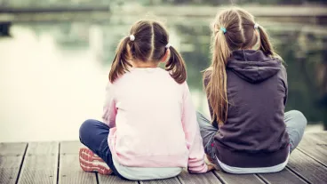 Two girls sitting on a wooden pier, their backs to the lens, surrounded by a serene waterbody.