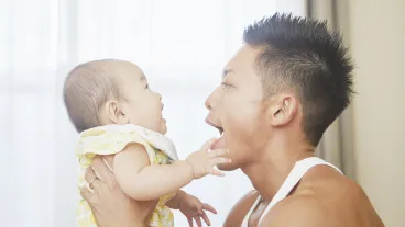 An adult holding a baby, with both facing each other indoors. The adult has their mouth open wide, and the baby is reaching toward their face.