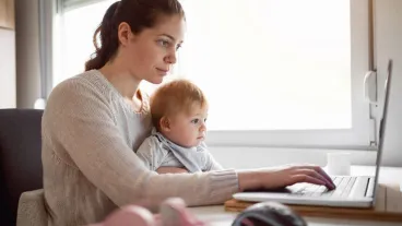 A woman works on a laptop at a desk while holding a baby on her lap. Both are looking at the computer screen.