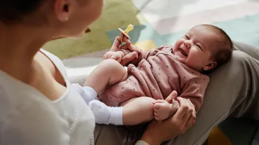 An adult holding a smiling baby dressed in a pink onesie, with the adult offering a pacifier.
