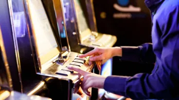 A person wearing a dark shirt presses buttons on the control panel of a slot machine in a casino.
