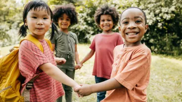 Four children stand outdoors holding hands and smiling at the camera, with trees and greenery in the background.