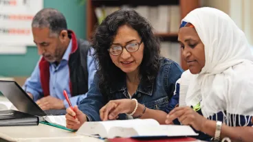 Three immigrant adults sit at a table studying; two women look at an open book together, while a man works on a laptop in the background.