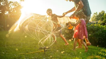 Father with children running through sprinkler at back yard in summer evening in sunset