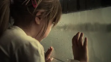A young child looks out a rain-covered window, touching the glass with one hand while raindrops are visible on the surface.