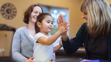 Preschool age girl high fives her proud, female physical therapist after completing a motor skills task in a health and wellness office.