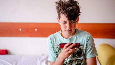 A young boy sits in his bedroom playing on a mobile device.
