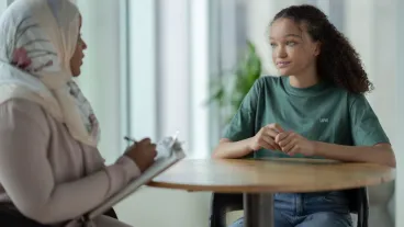 A young teen sits across the table from her therapist during a session together. She is dressed casually in a green t-shirt and has a neutral expression on her face as she answers her thera[ist's questions.