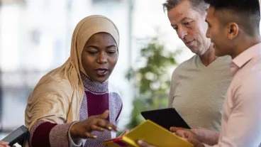Mixed ethnic group of business professionals at a networking conference/meeting room. They are standing looking at digital tablets together. Muslim woman leads the business meeting.