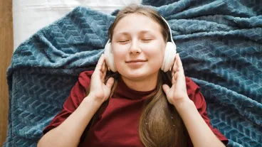 Young woman lying comfortably on a bed, eyes closed, wearing white headphones. She appears relaxed and content, enjoying her music in a cozy and peaceful bedroom setting.