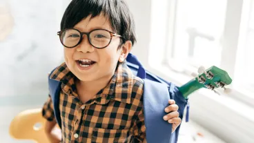 Young child wearing glasses and a plaid shirt smiles while carrying a blue backpack with part of a green ukulele visible inside.