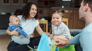 An adult reads a book to a smiling toddler while another adult holds a baby; they are sitting together on the floor in a home setting.
