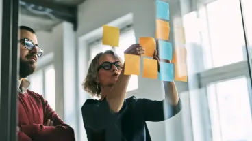 Two business professionals looking at sticky notes on a glass wall, engaged in a discussion and brainstorming new strategies for a project in an office.